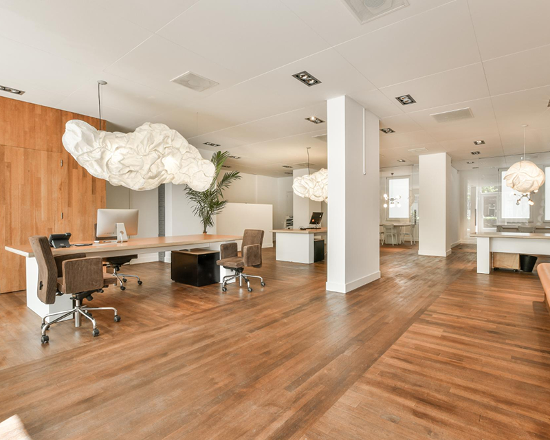 RCO-commercial-800x640 Modern open-plan commercial office with warm hardwood flooring, white support columns, sculptural cloud-shaped pendant lights above workstations, and desks with computers and rolling chairs.