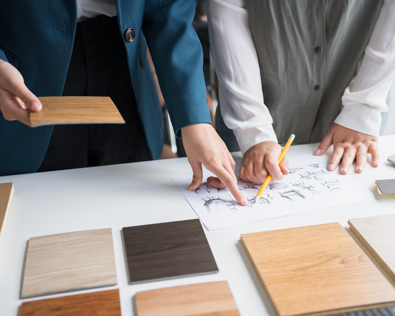 RCO-consultation-800x640 People reviewing furniture sketches and comparing wood flooring or laminate sample tiles in various shades on a table, with one holding a plank sample and another pointing at the drawings.