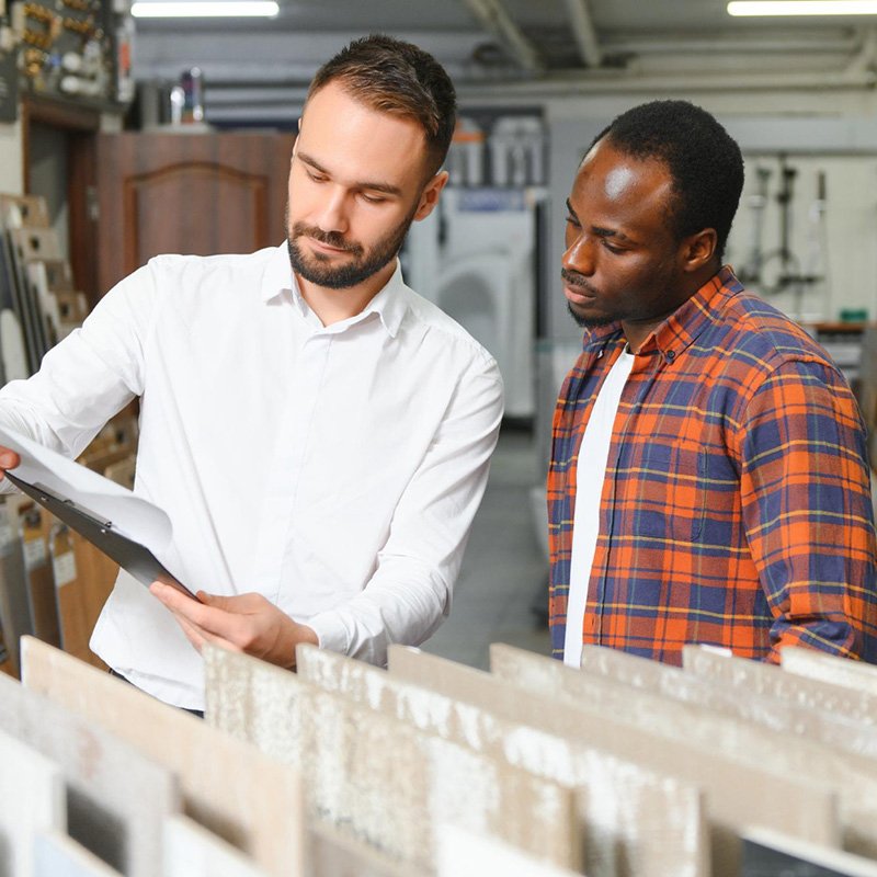 Flooring store associate holding a clipboard and reviewing tile or flooring samples with a customer in a showroom, both looking down at a display rack of material samples.