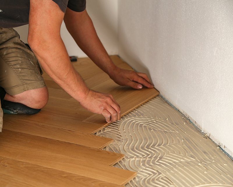 Flooring Installation Worker kneeling to press wood plank flooring into a layer of adhesive troweled onto the subfloor along the edge of a room.