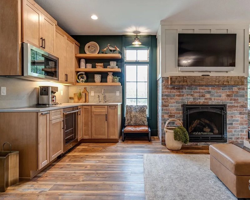 Residential Flooring Cozy living space with a wet bar featuring wood cabinetry and open shelving, dark green accent wall, and a brick fireplace with mounted TV, leather ottoman, and rug on wood flooring from Rafferty & Co.